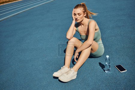 Foto de Joven deportista cansada en ropa deportiva sentada al lado de la botella de agua y el teléfono móvil después del entrenamiento - Imagen libre de derechos