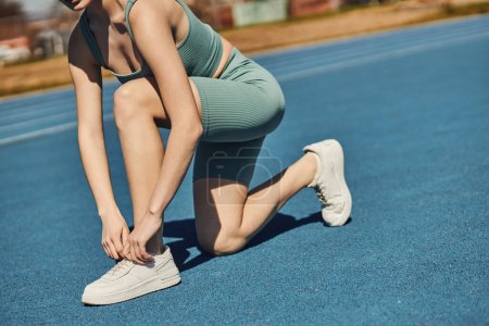 cropped sportswoman in activewear tying laces on white sneakers before running in jogging track