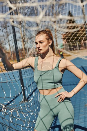 pretty sportswoman with ponytail standing in activewear standing with hand on hip near net outdoors