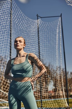 young woman with ponytail posing in sportswear near net while working out outdoors, sunny weather
