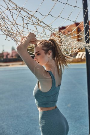 blonde junge Frau mit Pferdeschwanz posiert in Activwear, während sie nach dem Training in der Nähe des Netzes steht