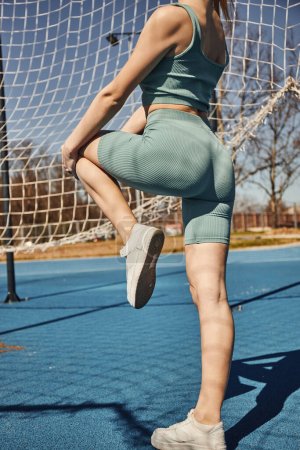 joven deportista recortada haciendo ejercicio en ropa deportiva y zapatillas de deporte cerca de la red al aire libre, fitness urbano