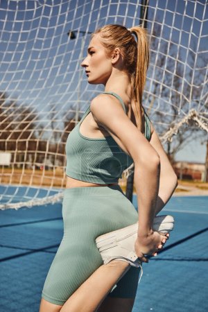 blonde junge Sportlerin mit Pferdeschwanz, die in Aktivkleidung in der Nähe des Netzes im Freien trainiert, das Bein streckt
