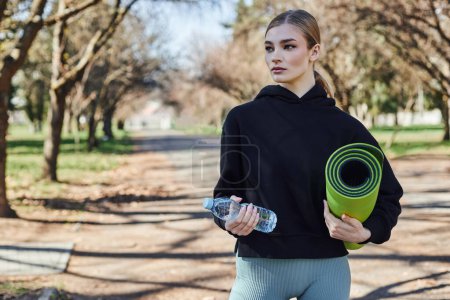 blonde and fit young woman in black sporty hoodie holding fitness mat and bottle of water in park