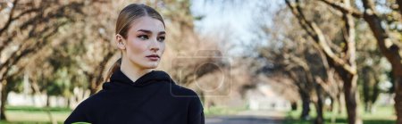 blonde and fit young woman in black sporty hoodie looking at camera after workout in park, banner