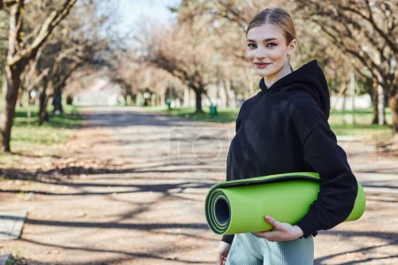 blonde and fit young woman in black sporty hoodie holding fitness mat after working out in park