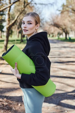pretty and fit young woman in black sporty hoodie holding fitness mat after working out in park