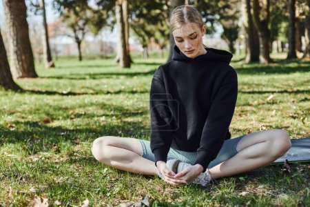 Foto de Mujer joven en forma con el pelo rubio y ropa deportiva sentado en la alfombra deportiva y meditando en el parque - Imagen libre de derechos