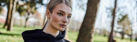 portrait of sporty young woman in black hoodie looking away in green park, nature and sport banner