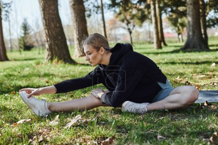 fit young woman with blonde hair and sportswear stretching while sitting on fitness mat in park