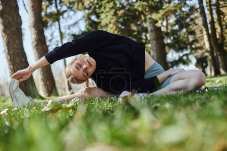 sporty young woman with blonde hair and sportswear stretching while sitting on green grass in park