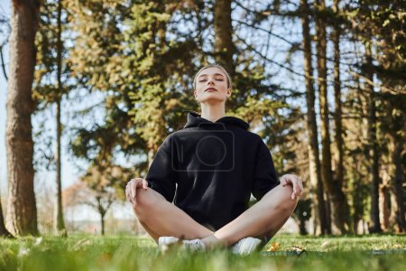 hübsche junge Frau mit blonden Haaren und Sportbekleidung sitzt auf Matte, während sie im Park meditiert