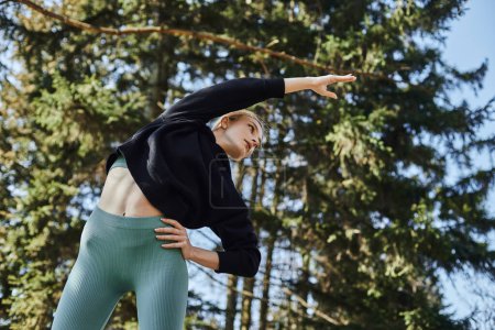 mujer joven y deportiva con cabello rubio y ropa deportiva estirando su cuerpo mientras hace ejercicio en el parque