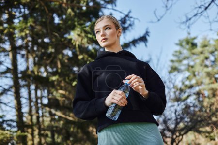fit young woman with blonde hair and sportswear holding bottle of water after working out in park