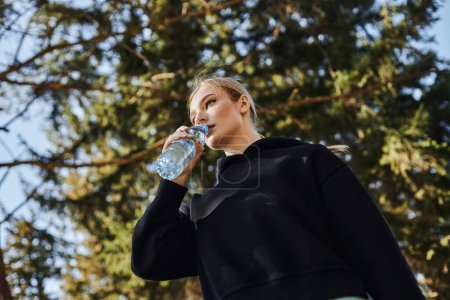 fit young woman with blonde hair and sportswear drinking fresh water after working out in park