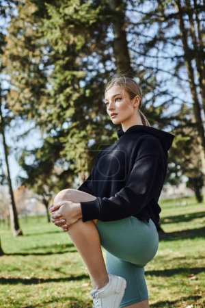 fit young woman with blonde hair and sportswear stretching leg while standing on grass in park