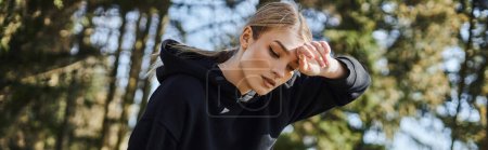 tired young woman with blonde hair and sportswear wiping sweat after working in green park, banner