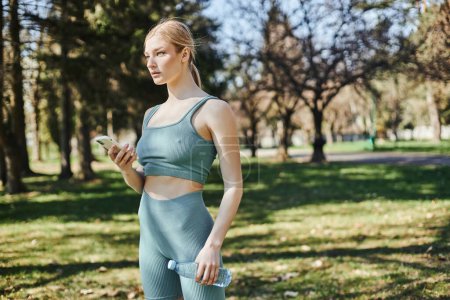 Blonde Sportlerin in aktiver Kleidung mit Wasserflasche und Smartphone im Park, Motivation und Sport