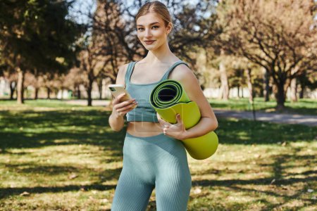 happy sportswoman in active wear holding smartphone and fitness mat while standing in park