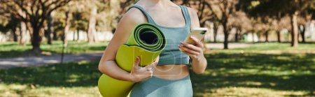 abgeschnittenes Banner einer Sportlerin in aktiver Kleidung mit Smartphone und Fitnessmatte im grünen Park