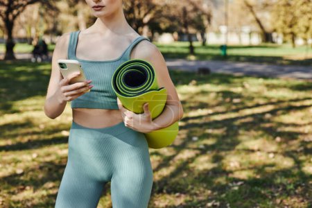 Ausgeschnittene Ansicht einer Sportlerin in aktiver Kleidung mit Smartphone und Fitnessmatte im grünen Park