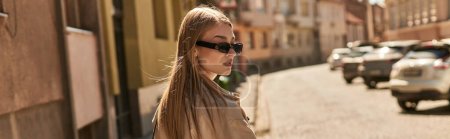 Blonde junge Frau in stylischer Sonnenbrille und beigem Trenchcoat spaziert durch die Stadt, urbanes Banner