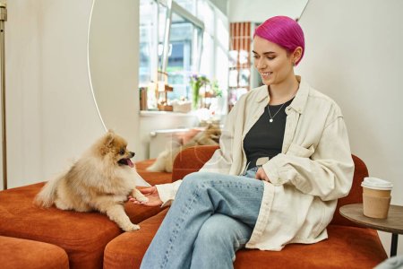 Foto de Alegre púrpura de pelo mujer sosteniendo pata de lindo pomeranian spitz cerca de café para ir en hotel de mascotas - Imagen libre de derechos