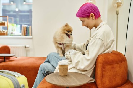 smiling purple-haired woman cuddling cute pomeranian spitz near coffee to go in lounge of dog hotel