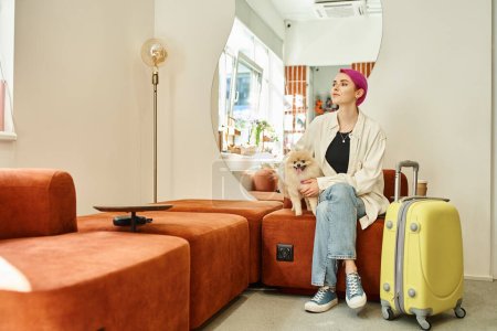 purple-haired woman with pomeranian spitz and suitcase waiting in lobby of pet hotel