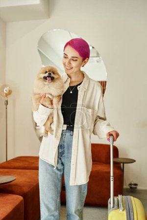 cheerful woman holding funny pomeranian spitz and travel bag in reception area of dog hotel