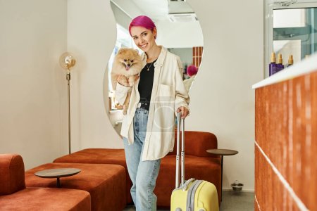 Foto de Alegre mujer de pelo morado mirando a la cámara mientras está de pie con perro peludo y maleta en el hotel de mascotas - Imagen libre de derechos
