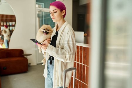woman with pomeranian spitz messaging on smartphone near reception desk and suitcase in dog hotel