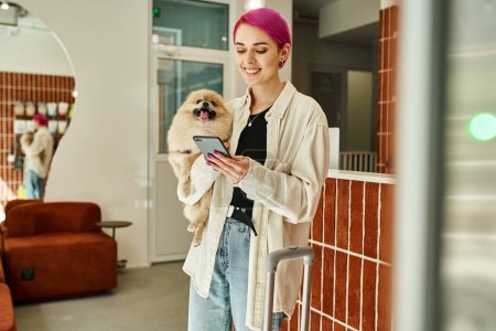 mujer alegre con pequeño perro esponjoso y teléfono inteligente cerca de recepción y bolsa de viaje en hotel perro