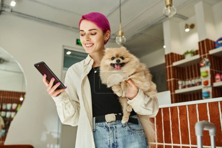 smiling woman with funny pomeranian spitz looking at mobile phone in reception area of pet hotel