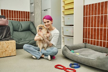 glücklicher Tiersitter umarmt Pommernspitz in der Nähe von weichem Hundebett und Spielzeug auf dem Fußboden im Haustierhotel