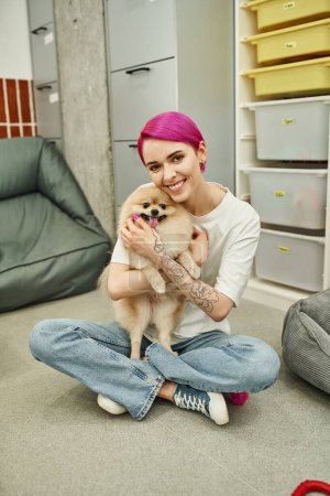 tätowierter Tiersitter mit lila Haaren umarmt flauschigen Hund und lächelt in die Kamera auf dem Fußboden im Haustierhotel