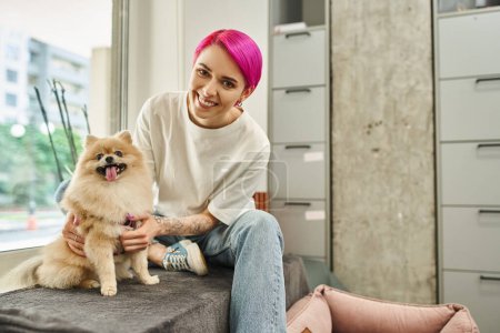fröhliche lila-haarige Hundesitter umarmt lustige Pommernspitze und lächelt in die Kamera im Haustierhotel