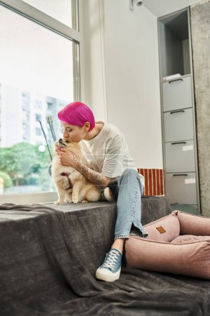 Téléchargez les photos : Travailleur de l'hôtel animal de compagnie attentionné avec les cheveux violets embrasser adorable spitz poméranien, chien amour et soins - en image libre de droit