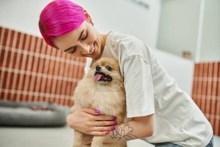 lächelnde Hundesitterin mit lila Haaren kuschelt süße Pommernspitze im Haustierhotel, liebevolle Pflege