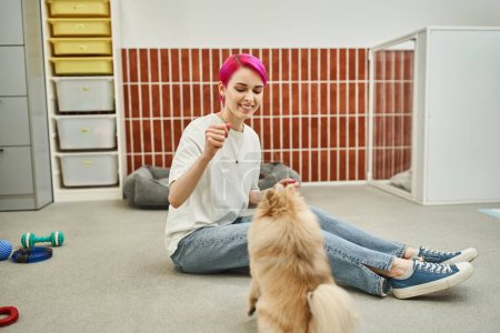 smiley dog caregiver training pomeranian spitz while sitting on floor and holding treat in hand