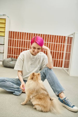 happy woman with purple hair sitting on floor near pomeranian spitz in pet hotel, loving dog sitter