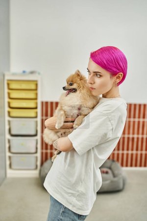 Aufmerksame Tierpflegerin mit lila Haaren hält Pommernspitz in der Hand und schaut im Hundehotel weg