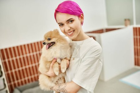 smiling female pet sitter with purple hair holding pomeranian spitz in pet hotel, care and bonding