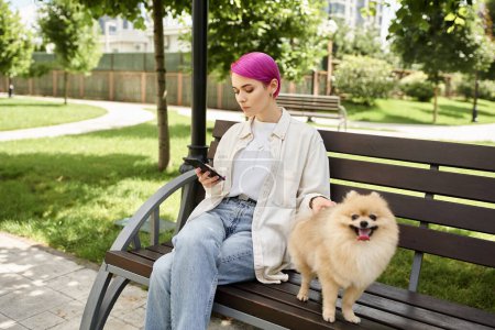 púrpura mujer de pelo en red en el teléfono inteligente mientras descansa en el banco del parque cerca de spitz pomeranian lindo