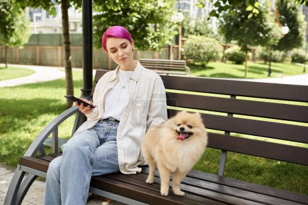 Foto de Divertido pomeranian spitz de pie en el banco cerca de elegante mujer de pelo morado con teléfono inteligente - Imagen libre de derechos