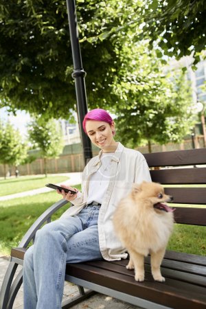 Foto de Alegre mujer de pelo morado con teléfono inteligente mirando spitz pomeraniano adorable en el banco en el parque - Imagen libre de derechos