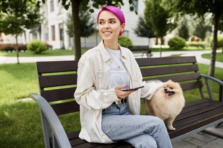 Foto de Alegre mujer de pelo morado con teléfono inteligente mirando hacia otro lado mientras está sentado en el banco del parque cerca del perro - Imagen libre de derechos
