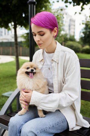 Foto de Elegante mujer de pelo morado abrazando spitz pomeraniano adorable mientras está sentado en el banco en el parque - Imagen libre de derechos