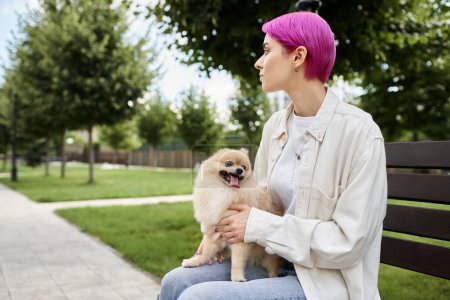 mujer de pelo púrpura soñadora con esponjoso spitz pomeraniano sentado en el banco en el parque y mirando hacia otro lado