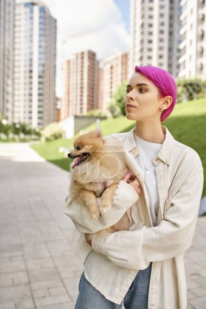 purple-haired woman looking away while walking on city street with pomeranian spitz in hands